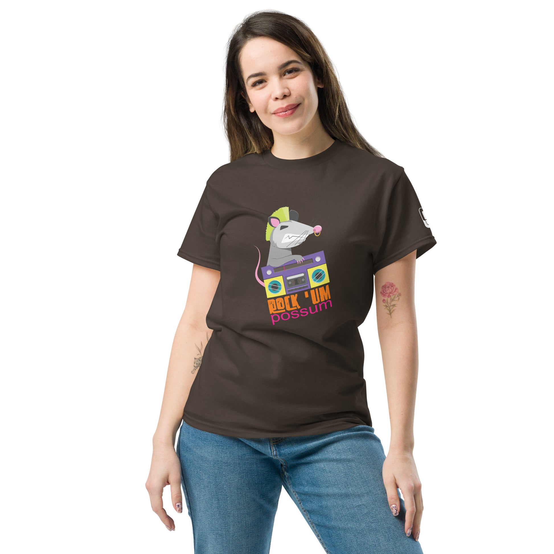A young Caucasian woman with shoulder-length dark hair, smiling subtly while standing casually. She is wearing a brown t-shirt featuring a fun graphic of a possum with a neon green mohawk, operating a purple boombox. The text "ROCK 'UM POSSUM" in colorful, playful lettering enhances the quirky charm of the shirt. Her relaxed pose and light denim jeans complement the laid-back vibe of the t-shirt design.