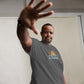 A middle-aged African American man poses confidently with his hand raised towards the camera, giving a stop gesture. He wears a charcoal gray t-shirt with a colorful design featuring stylized mountains in yellow and orange. Below the the graphic is "HAPPY CAMPER" in pale blue. His expression is serious yet friendly. The setting is a softly lit indoor environment, enhancing the focus on his expressive pose and the detailed graphic on his shirt.
