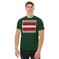 A young man with short, light brown hair is wearing a dark green t-shirt featuring a festive, Christmas-themed graphic. The shirt displays a large rectangular print with holiday patterns in red, black, and green, similar to a traditional holiday sweater design. 'TIS THE SEASON' is prominently included in the design, written in green against a white background, enhancing the festive mood.