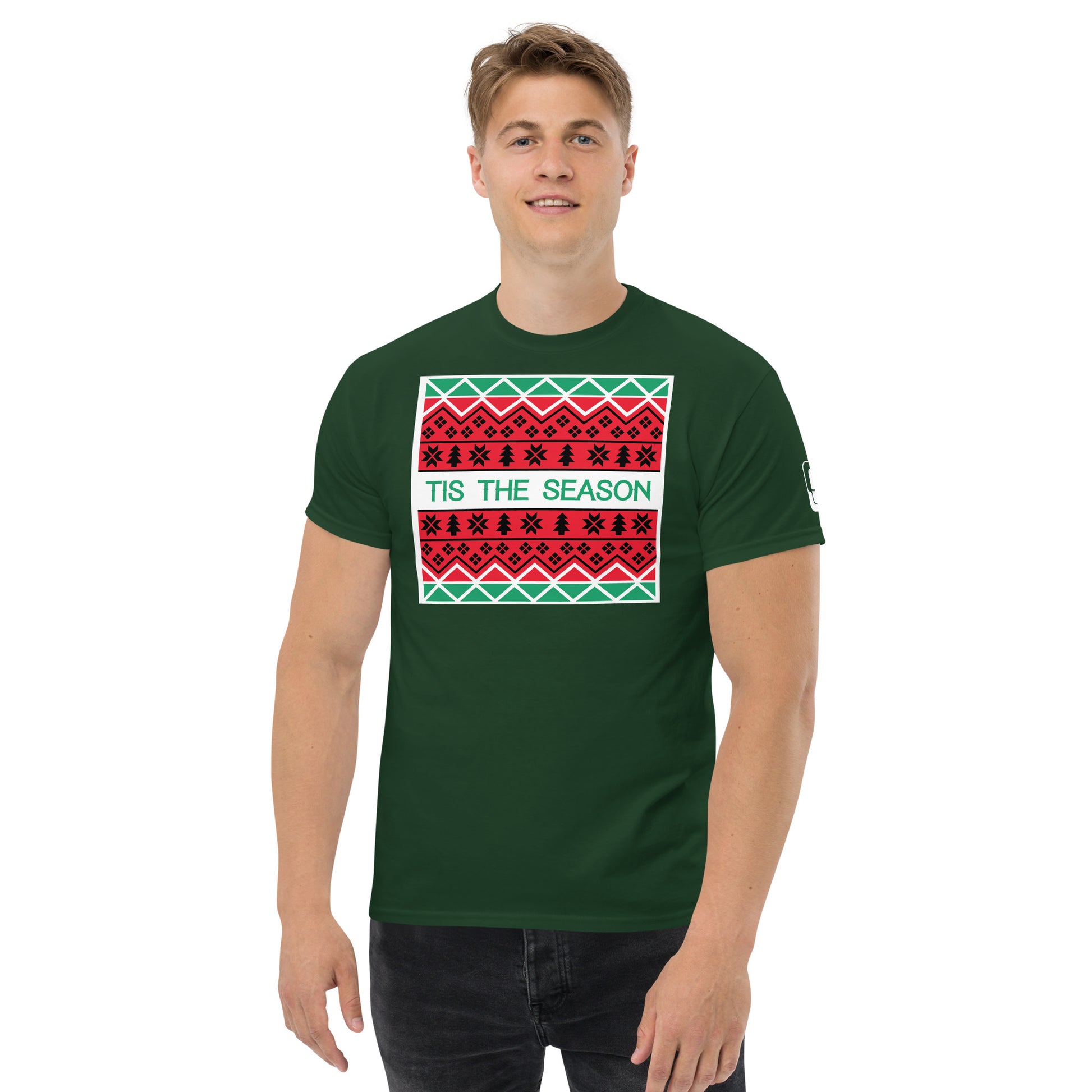 A young man with short, light brown hair is wearing a dark green t-shirt featuring a festive, Christmas-themed graphic. The shirt displays a large rectangular print with holiday patterns in red, black, and green, similar to a traditional holiday sweater design. 'TIS THE SEASON' is prominently included in the design, written in green against a white background, enhancing the festive mood.
