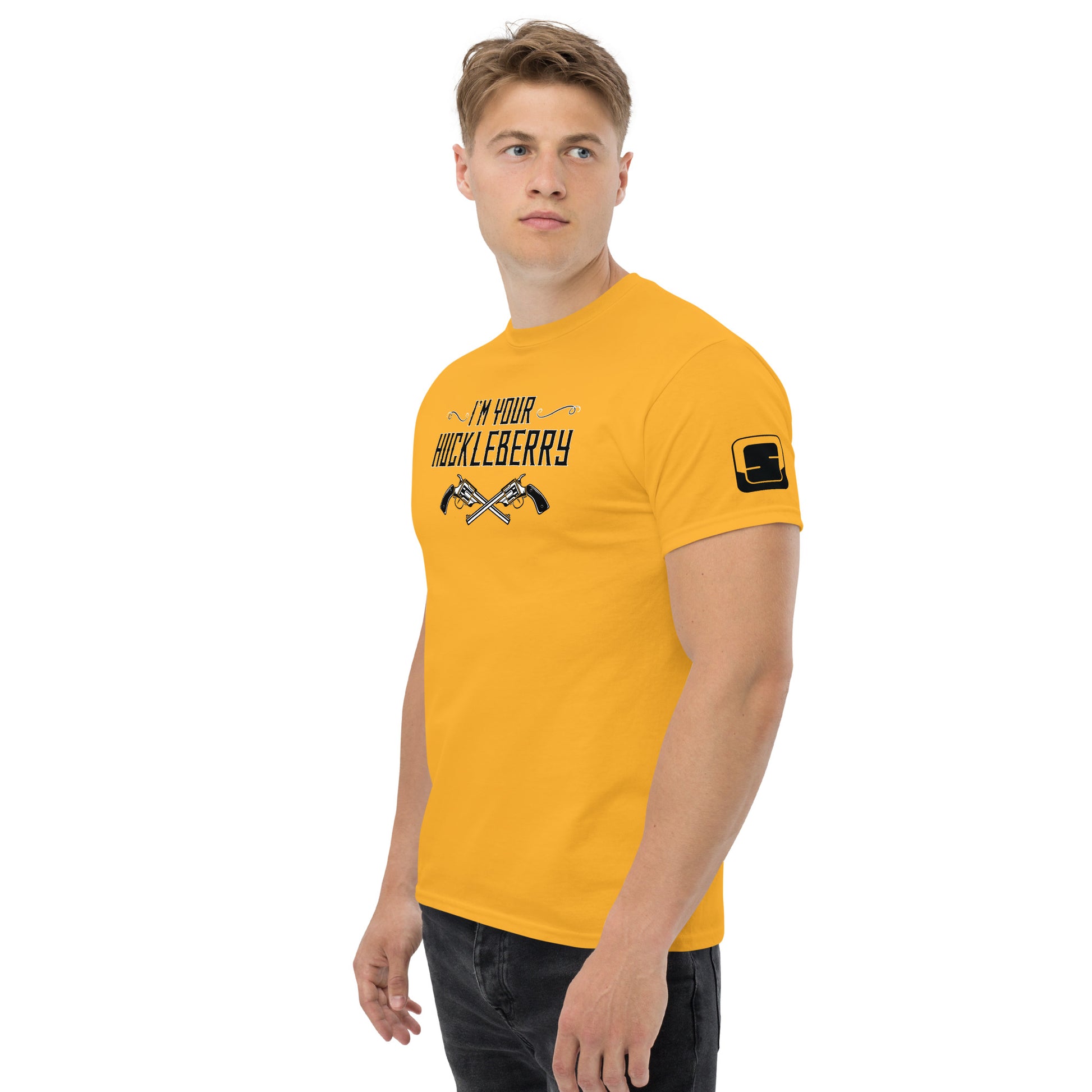 The image features a young man in a mustard yellow t-shirt that reads "I'm Your Huckleberry" in a bold, Western-style font with two crossed revolvers depicted beneath the text. The design is in black, creating a striking contrast against the yellow fabric. The man stands in a casual pose with a slight turn to his left, looking directly at the camera with a neutral expression. A small logo patch is visible on the sleeve of the t-shirt, adding a modern touch to the vintage-inspired design.