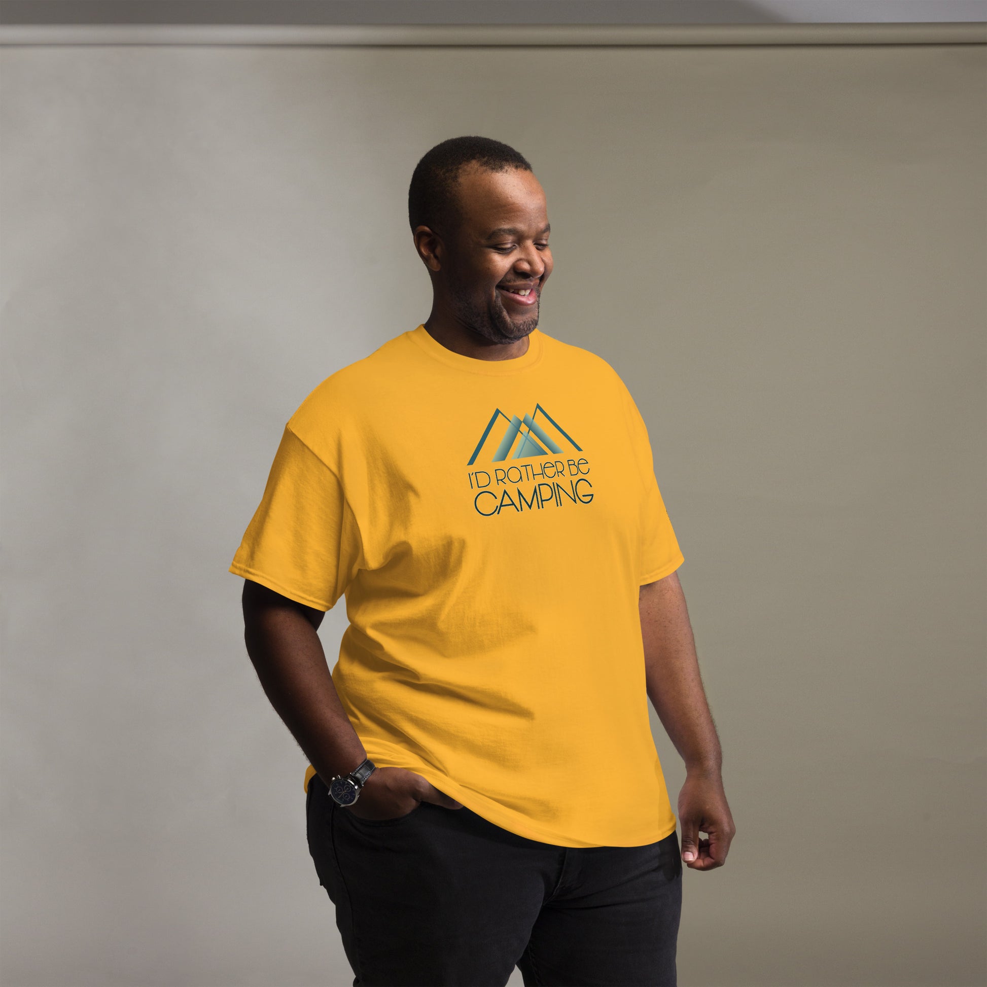 A cheerful African American man wearing a golden yellow t-shirt printed with "I'D RATHER BE CAMPING" and a stylized mountain graphic in teal. He is casually dressed, smiling, and looking downwards to his right, conveying a relaxed and happy demeanor. The backdrop is a soft grey, highlighting his bright shirt and joyful expression.