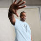 An African American man in a light blue T-shirt featuring a geometric deer head design extends his hand towards the camera in a stopping gesture. He looks directly at the viewer with a serious expression, standing against a light gray backdrop. His stance conveys a dynamic and engaging presence.