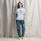 A woman stands confidently in a studio setting, wearing a light-blue t-shirt with the phrase 'CAMP MORE' and a stylized graphic of mountains in orange. She pairs the shirt with loose-fitting blue jeans and white sneakers. The backdrop is a neutral toned draped fabric, adding a soft texture to the scene.