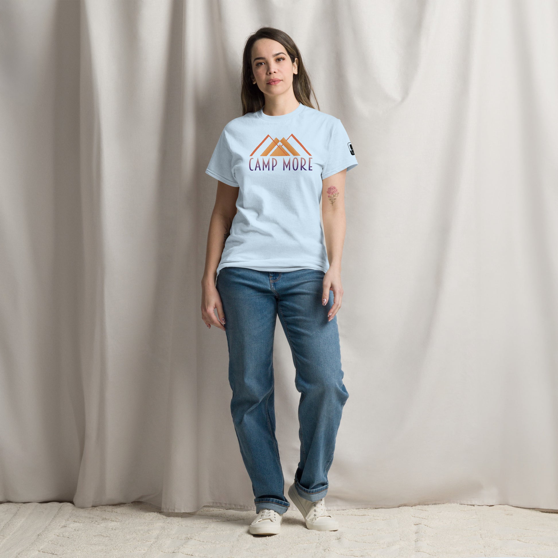 A woman stands confidently in a studio setting, wearing a light-blue t-shirt with the phrase 'CAMP MORE' and a stylized graphic of mountains in orange. She pairs the shirt with loose-fitting blue jeans and white sneakers. The backdrop is a neutral toned draped fabric, adding a soft texture to the scene.