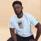 A young African American man with short black hair and glasses models a white T-shirt featuring a vibrant Independence Day graphic. The design includes the Statue of Liberty and an American flag, surrounded by colorful fireworks in red, blue, and purple bursts. The number '1776' prominently displayed at the top commemorates the year of American independence. He is seated casually, leaning slightly forward, giving a subtle, confident look to the camera against a soft beige background.