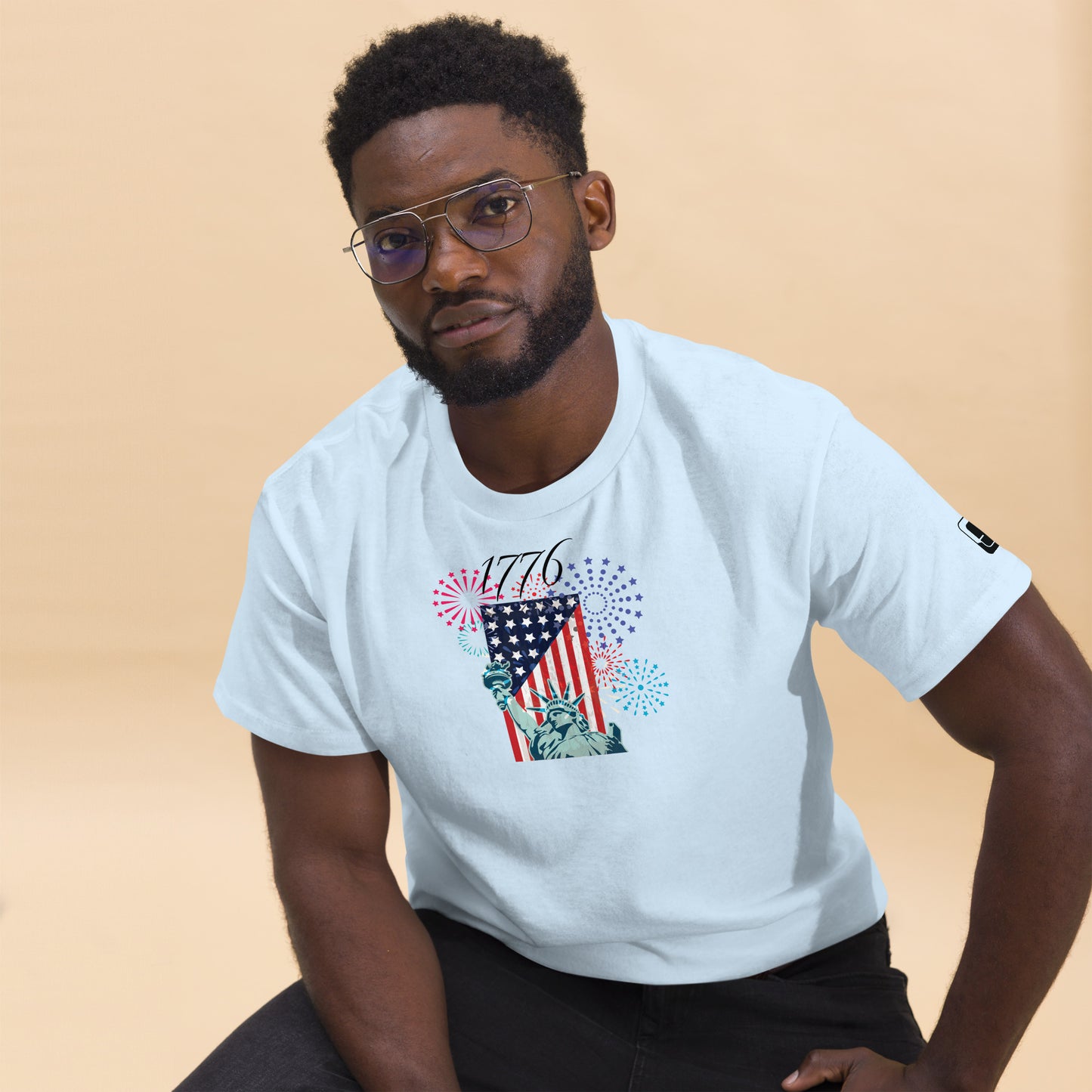 A young African American man with short black hair and glasses models a white T-shirt featuring a vibrant Independence Day graphic. The design includes the Statue of Liberty and an American flag, surrounded by colorful fireworks in red, blue, and purple bursts. The number '1776' prominently displayed at the top commemorates the year of American independence. He is seated casually, leaning slightly forward, giving a subtle, confident look to the camera against a soft beige background.