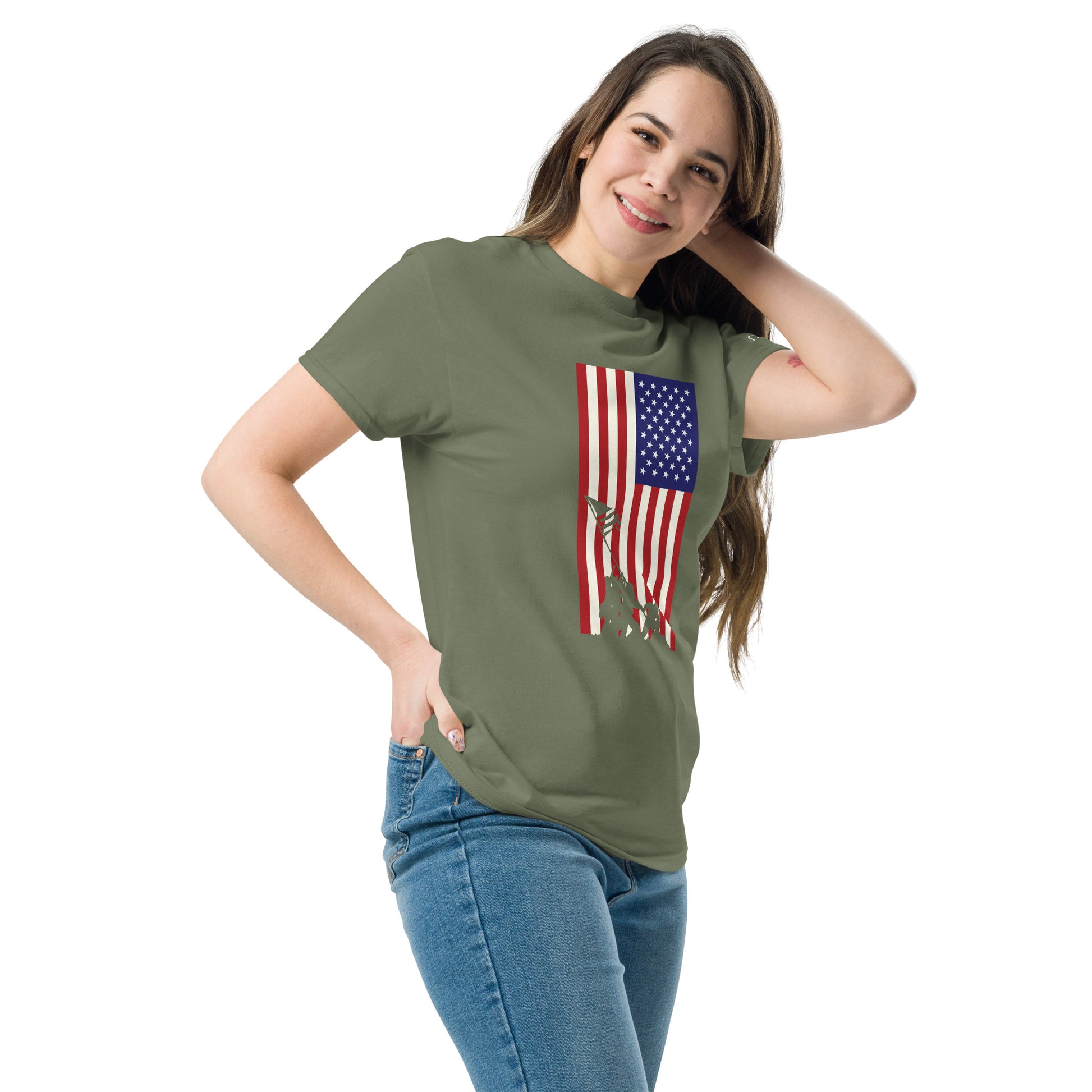 A young woman with long brown hair poses casually in a fitted olive green T-shirt adorned with a patriotic American flag design. The flag features stars and stripes, with a silhouette of the iconic Iwo Jima flag-raising scene, adding a historical and proud tone to the shirt. She stands smiling gently against a plain background, highlighting the tee's graphics.