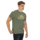 A young Caucasian man stands casually, wearing a muted green t-shirt that features a stylized mountain design in yellow, and orange orange. Below the graphic, the phrase "HAPPY CAMPER" is prominently displayed in pale blue letters. His expression is serious and contemplative as he looks off to the side, dressed in dark jeans and standing against a plain background, which highlights the vivid design on his shirt. His overall demeanor is relaxed yet attentive.