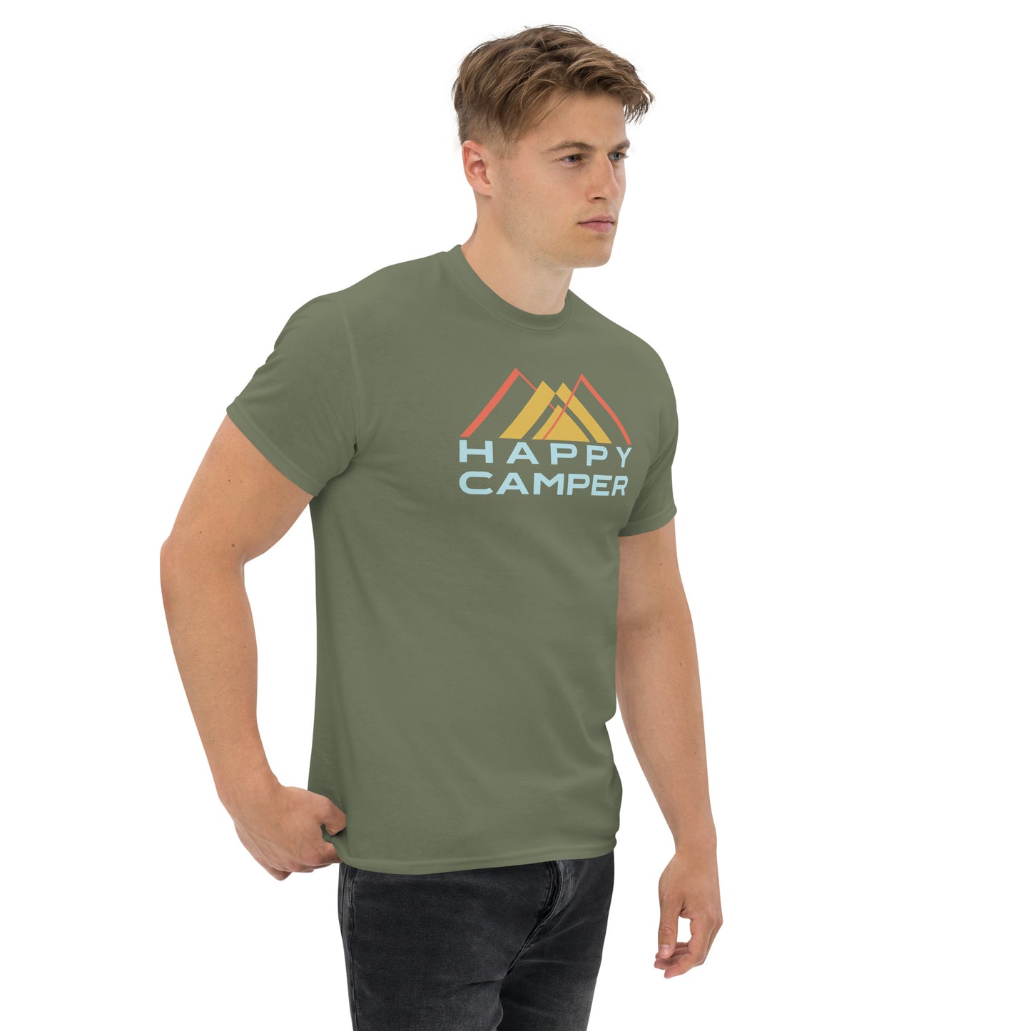 A young Caucasian man stands casually, wearing a muted green t-shirt that features a stylized mountain design in yellow, and orange orange. Below the graphic, the phrase "HAPPY CAMPER" is prominently displayed in pale blue letters. His expression is serious and contemplative as he looks off to the side, dressed in dark jeans and standing against a plain background, which highlights the vivid design on his shirt. His overall demeanor is relaxed yet attentive.