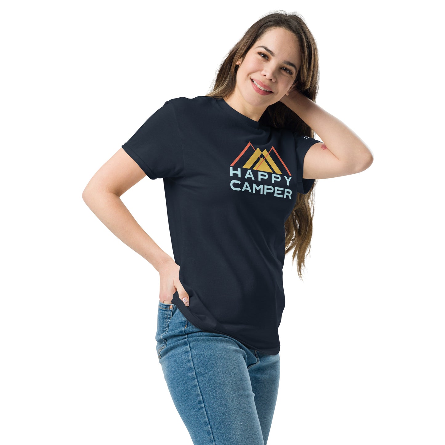 A young Caucasian woman poses playfully, her left hand on her hip and right hand touching her hair, smiling gently towards the camera. She wears a navy blue t-shirt with a colorful  design, featuring stylized mountains in shades of orange, and yellow, with the phrase "HAPPY CAMPER" displayed prominently in a clean, pale blue font. She pairs the shirt with classic blue jeans and stands against a plain background, highlighting her casual yet stylish look.