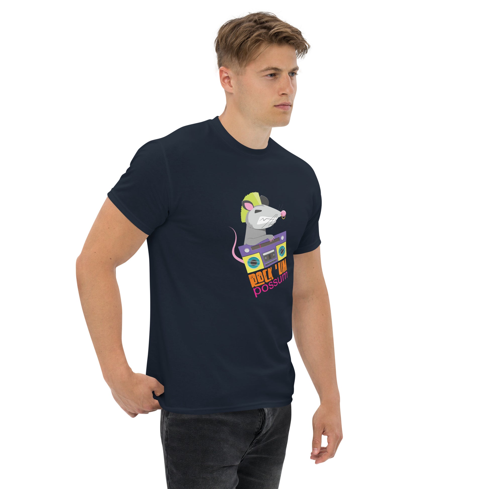 A young Caucasian man with short blonde hair wears a dark navy blue t-shirt featuring a playful graphic of a possum with a neon green mohawk, holding a colorful purple boombox. The graphic, which includes the fun phrase "ROCK 'UM POSSUM" in bold, multicolored letters beneath the image, adds a vibrant, quirky touch to the t-shirt. The man stands casually with a slight turn to his left, giving a subtle, focused expression.