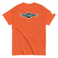 A plain orange-colored t-shirt laid flat, featuring the Shapetrix Entertainment logo centrally placed. The logo includes the brand name in stylish teal and white lettering inside a black circle, accented by elongated white wing-like graphics on both sides. A tag on the left sleeve shows a tiny black and white Shapetrix Entertainment label, adding a sophisticated touch to the vibrant shirt.