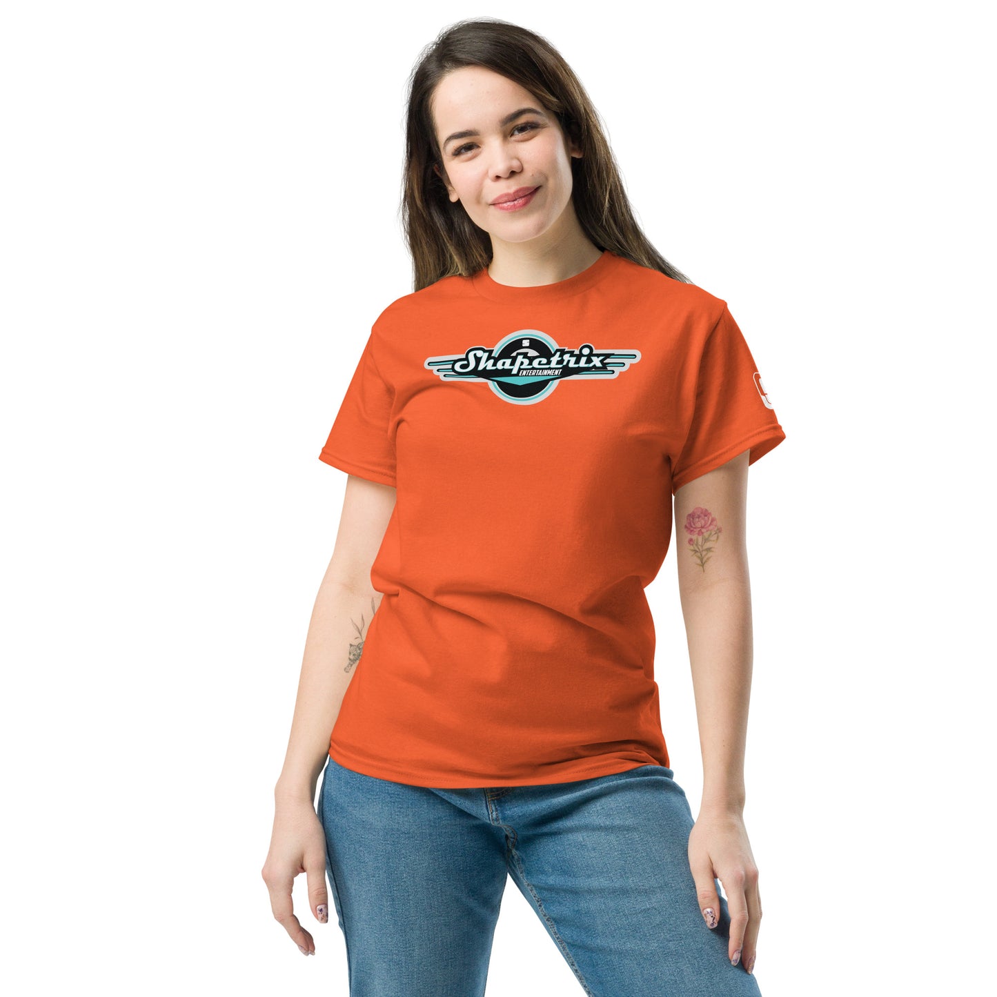 A young Caucasian woman with shoulder-length hair, casually standing with a slight turn to her right, wearing a terracotta-colored t-shirt. The t-shirt features the Shapetrix Entertainment logo, prominently displayed on the chest. The logo combines teal and white lettering with retro-style wings and a circular black background, creating a vintage-modern aesthetic. The relaxed fit of the t-shirt complements her casual denim jeans, adding to the overall laid-back and approachable vibe.