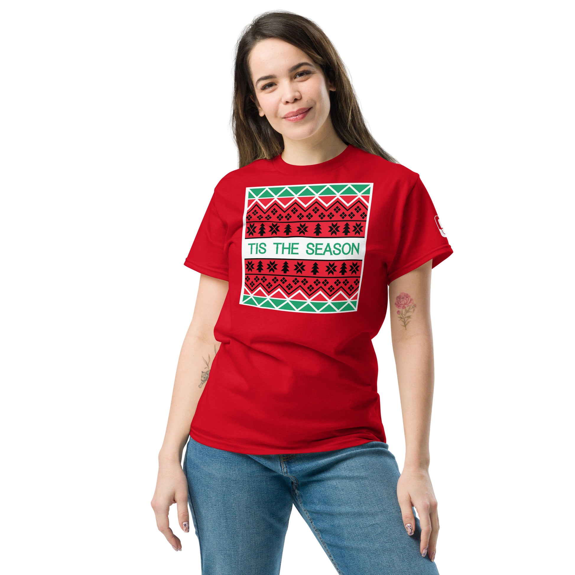 A woman stands casually with one hand on her hip, wearing a vibrant red t-shirt featuring a festive, Christmas-themed graphic print. The print includes a pattern similar to traditional holiday sweaters with rows of black and white symbols like stars and geometric designs on a green background. Centered in a white text box is the phrase 'TIS THE SEASON', adding to the cheerful holiday spirit of the shirt.