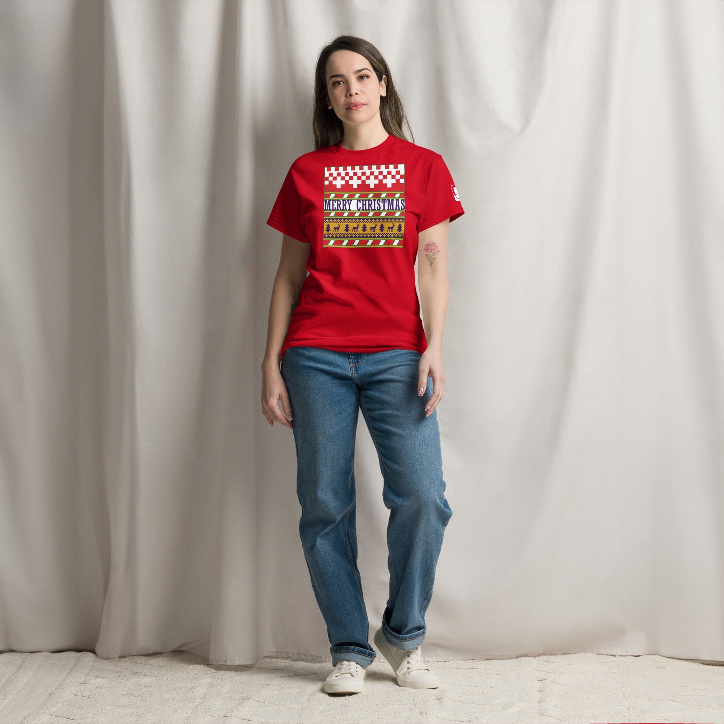 A woman stands confidently in a studio setting, wearing a bright red t-shirt with a festive 'Merry Christmas' design. The shirt is styled with a red and white checkerboard pattern on the top, a green stripe, and a yellow band featuring reindeer and trees. She pairs her holiday-themed tee with casual blue jeans and white sneakers, presenting a relaxed yet festive look.
