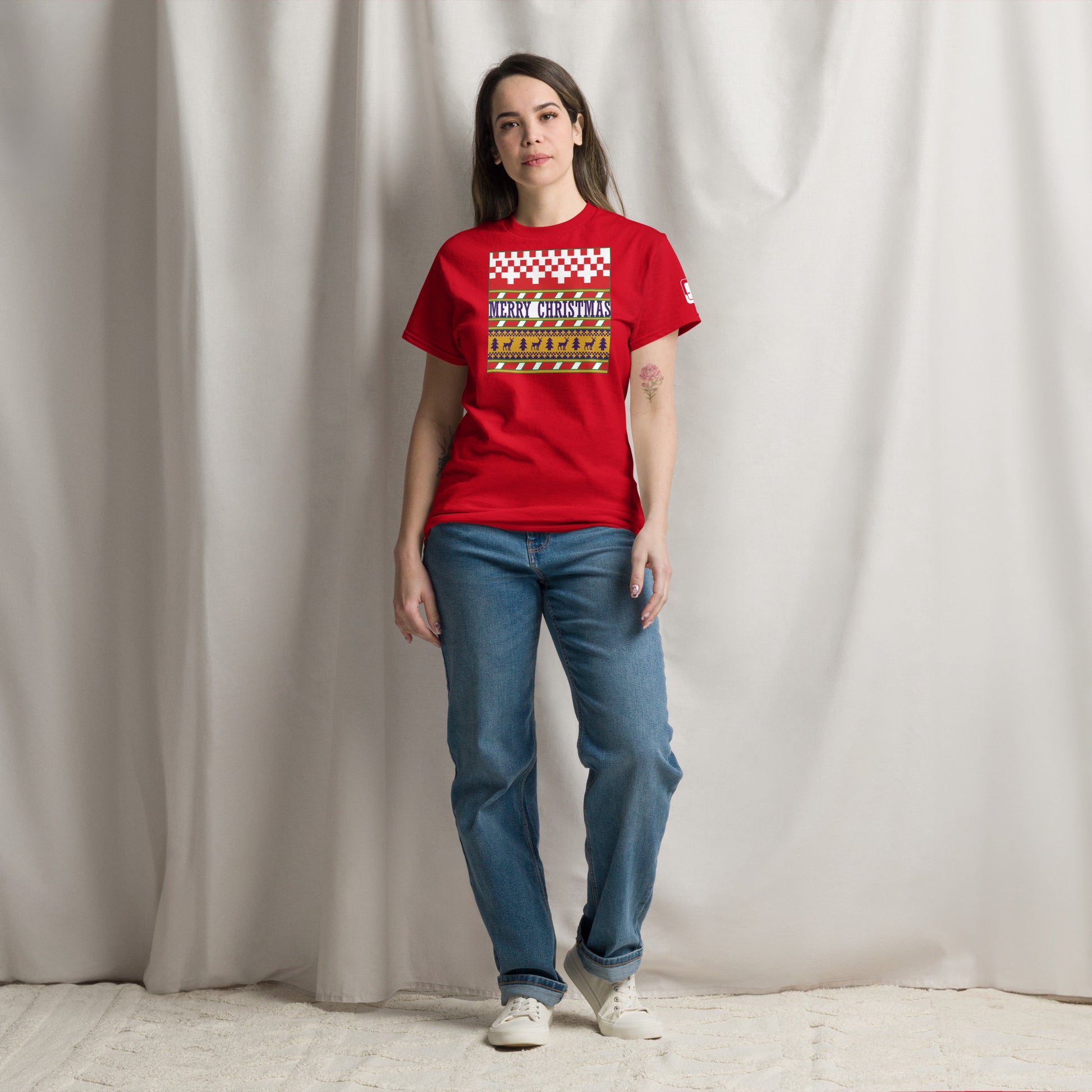 A woman stands confidently in a studio setting, wearing a bright red t-shirt with a festive 'Merry Christmas' design. The shirt is styled with a red and white checkerboard pattern on the top, a green stripe, and a yellow band featuring reindeer and trees. She pairs her holiday-themed tee with casual blue jeans and white sneakers, presenting a relaxed yet festive look.