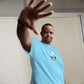 Man wearing a light blue short-sleeved t-shirt with a 'Tee Time' graphic showing a golf ball character on grass on the chest standing against a plain beige backdrop with an out stitched arm.