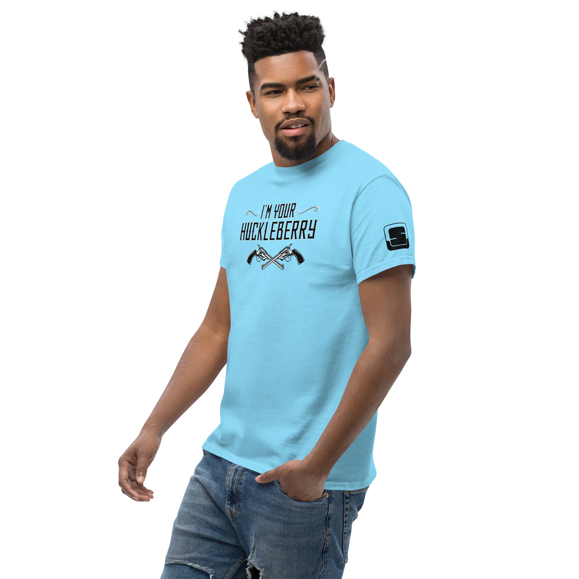 The image features a stylish African American man wearing a light blue t-shirt that says "I'm Your Huckleberry" in a vintage Western font with an illustration of two crossed revolvers beneath the text. The design is in black, providing a striking contrast against the light blue background of the shirt. He is looking confidently towards the camera with a friendly, engaging smile. His stance is relaxed, with one hand partially tucked into the pocket of his torn jeans. A small logo patch is visible o