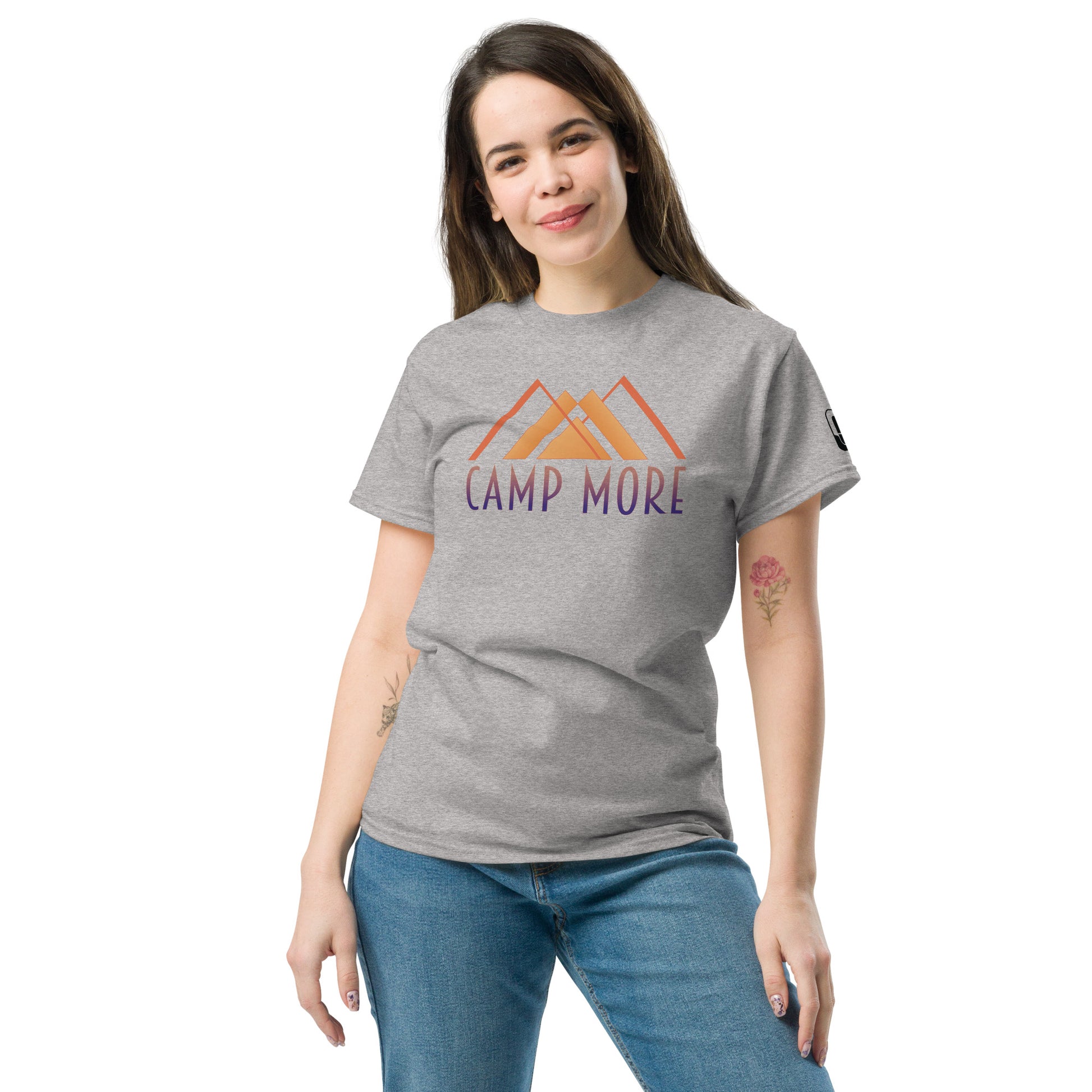 A woman poses casually in a studio setting, wearing a light gray t-shirt emblazoned with the phrase 'CAMP MORE' in bold orange and purple, accompanied by an abstract illustration of mountains. She complements the t-shirt with fitted blue jeans and has visible tattoos on her arms. The light, neutral background focuses attention on her relaxed and friendly demeanor.