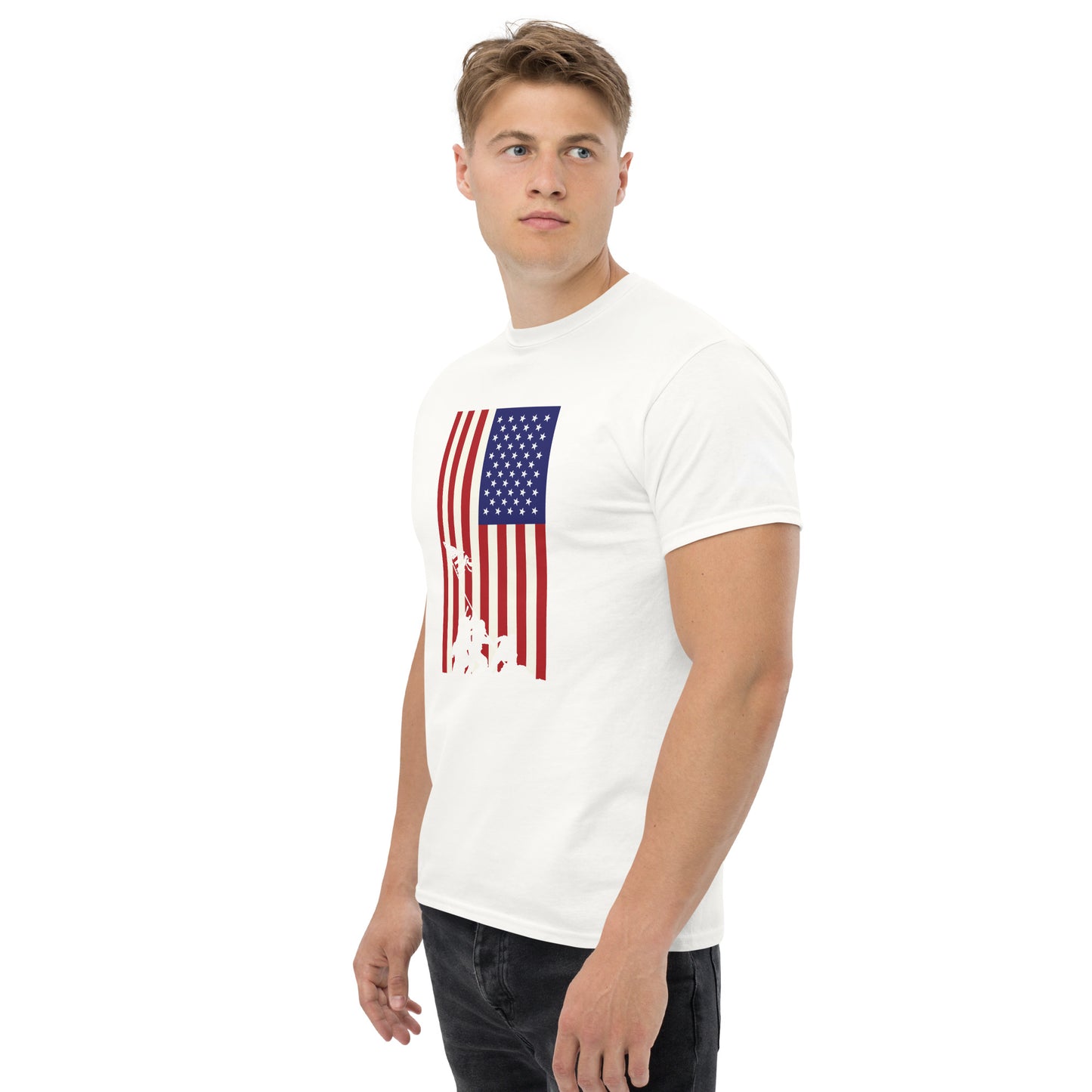 A young Caucasian man in a plain white T-shirt featuring a distressed American flag print with an Iwo Jima silhouette at the bottom stands confidently. The flag design includes a blue square with white stars on the upper left, merging into red and white stripes that flow into the silhouette. He models the shirt in a simple stance, looking slightly to the side, against a neutral background that emphasizes the bold, historical-themed shirt design.