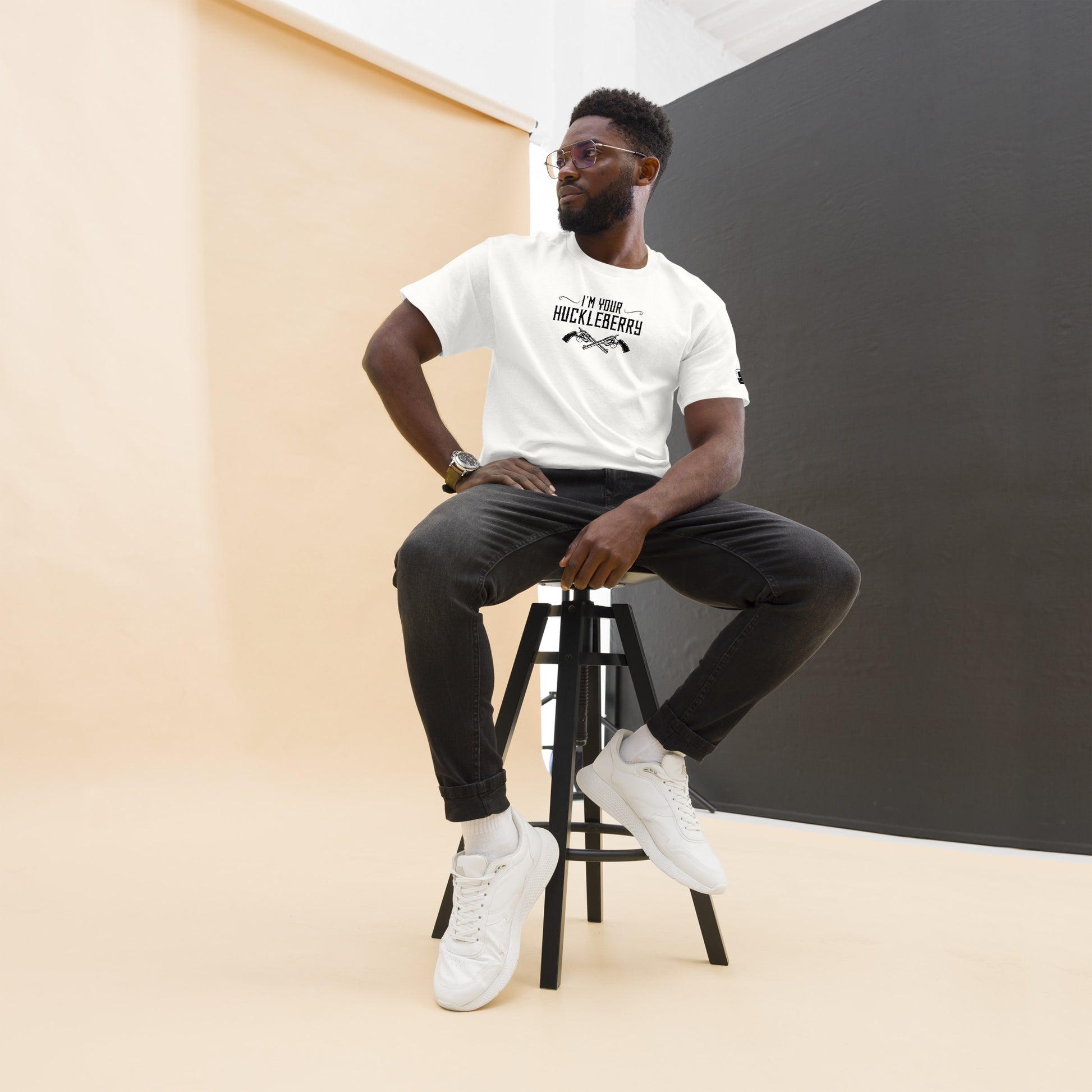 The image shows an African American man in a fashionable pose, seated on a stool. He is wearing a white t-shirt with the phrase "I'm Your Huckleberry" in a bold, black font, accompanied by two crossed revolvers. He pairs this with dark jeans and white sneakers. His look is completed with a stylish pair of glasses and a wristwatch, adding a touch of sophistication. The background alternates between a warm peach and a neutral gray, providing a modern, minimalist aesthetic that highlights his attire.