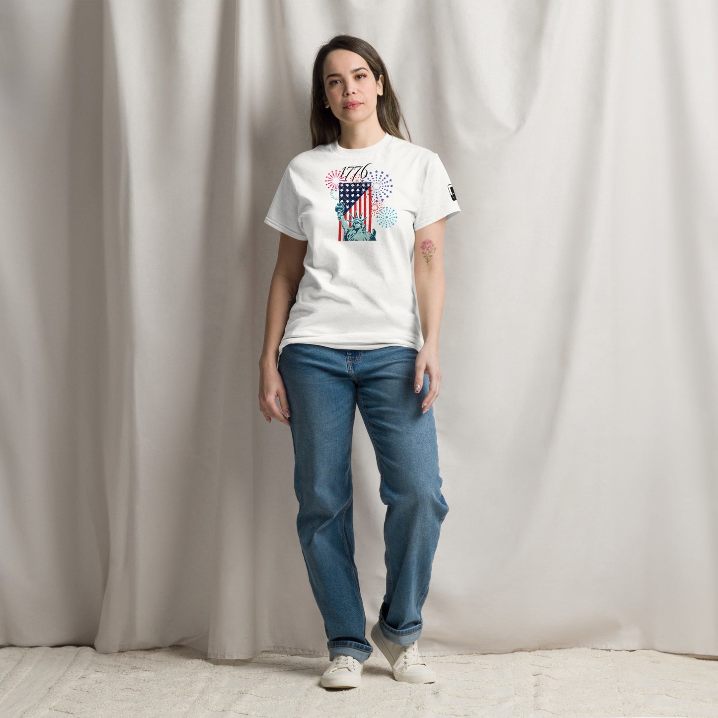 A young Caucasian woman with long, straight brown hair stands casually in a studio, modeling a white T-shirt adorned with a vibrant graphic celebrating American independence. The design features a striking American flag, the Statue of Liberty, and colorful fireworks, complemented by the year '1776' in bold lettering. She wears loose blue jeans and white sneakers and a relaxed, subtle smile against