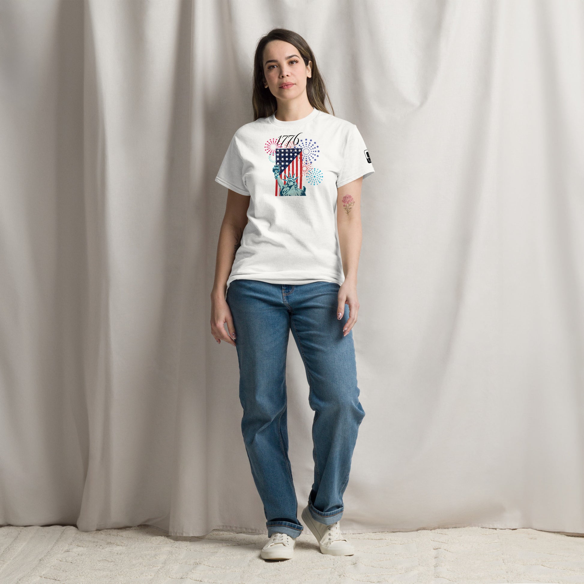 A young Caucasian woman with long, straight brown hair stands casually in a studio, modeling a white T-shirt adorned with a vibrant graphic celebrating American independence. The design features a striking American flag, the Statue of Liberty, and colorful fireworks, complemented by the year '1776' in bold lettering. She wears loose blue jeans and white sneakers and a relaxed, subtle smile against
