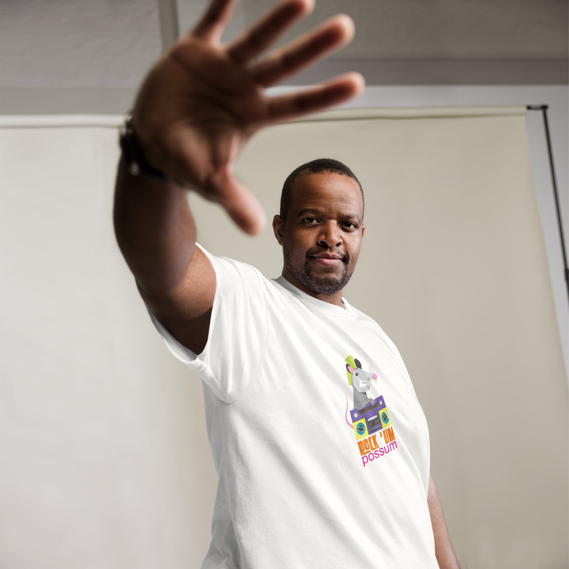 A middle-aged African American man in a white t-shirt reaches towards the camera with an open hand, partially obscuring his face in a playful gesture. His t-shirt features a whimsical graphic of a possum with a neon green mohawk, cheerfully operating a purple boombox, accompanied by the text "ROCK 'UM POSSUM" in vibrant, colorful letters. His relaxed pose and subtle smile convey a casual, friendly demeanor.