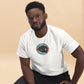 An African American man in stylish eyeglasses poses casually while wearing a white t-shirt that features a central graphic of a poker chip enclosing a royal flush poker hand. The chip's design includes red and gray accents with the text "Royal Flush" at the top. He pairs the tee with black trousers, sitting against a neutral beige background that emphasizes the t-shirt's graphic, projecting a relaxed yet attentive demeanor.