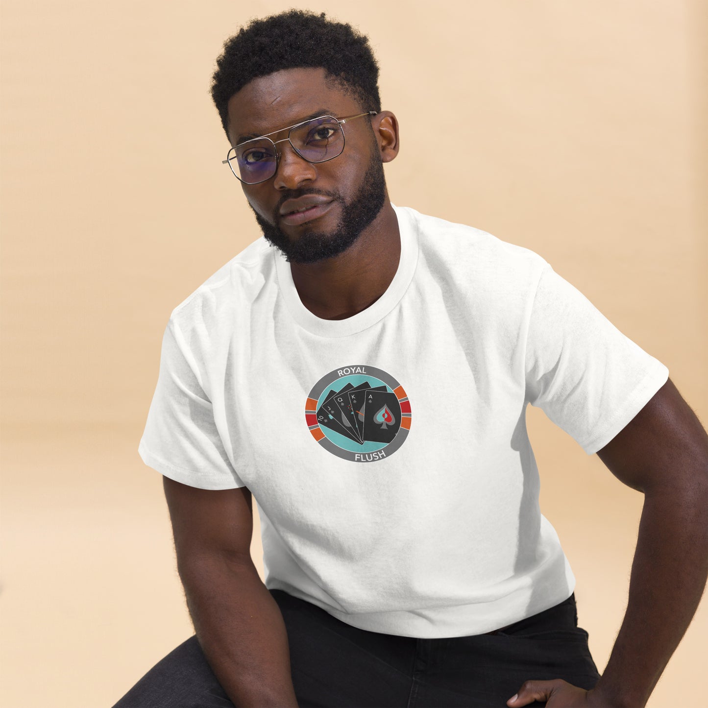 An African American man in stylish eyeglasses poses casually while wearing a white t-shirt that features a central graphic of a poker chip enclosing a royal flush poker hand. The chip's design includes red and gray accents with the text "Royal Flush" at the top. He pairs the tee with black trousers, sitting against a neutral beige background that emphasizes the t-shirt's graphic, projecting a relaxed yet attentive demeanor.