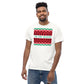 A cheerful man with a bright smile models a white t-shirt adorned with a festive, Christmas-themed graphic print. The print includes a white rectangle featuring three rows of traditional holiday patterns in red, black, and green, reminiscent of a holiday sweater. The phrase 'TIS THE SEASON' is prominently displayed in green, adding a joyful touch to the seasonal attire.