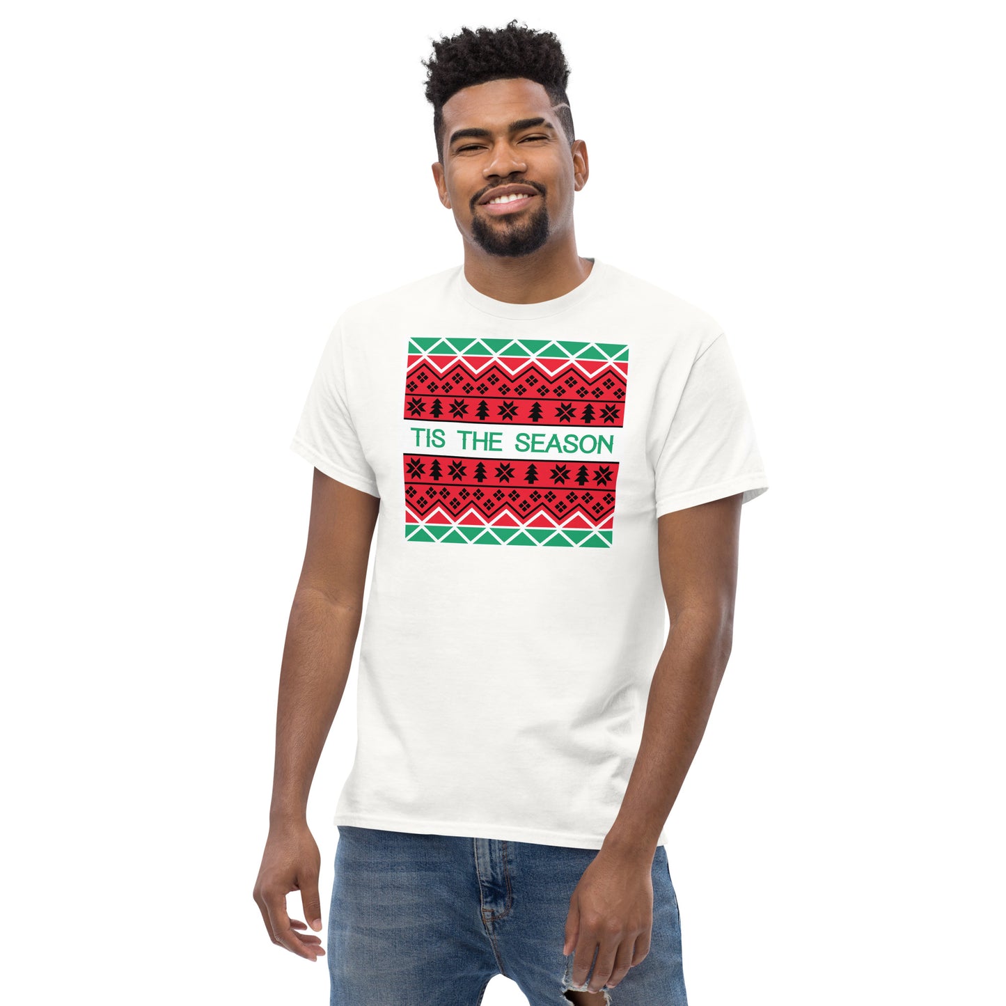 A cheerful man with a bright smile models a white t-shirt adorned with a festive, Christmas-themed graphic print. The print includes a white rectangle featuring three rows of traditional holiday patterns in red, black, and green, reminiscent of a holiday sweater. The phrase 'TIS THE SEASON' is prominently displayed in green, adding a joyful touch to the seasonal attire.