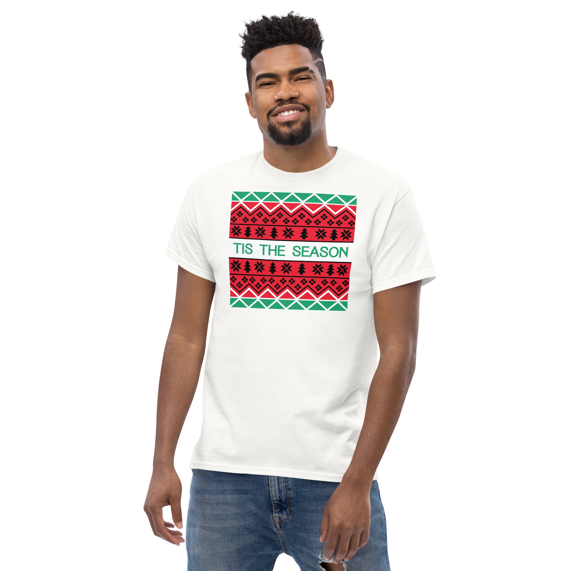 A cheerful man with a bright smile models a white t-shirt adorned with a festive, Christmas-themed graphic print. The print includes a white rectangle featuring three rows of traditional holiday patterns in red, black, and green, reminiscent of a holiday sweater. The phrase 'TIS THE SEASON' is prominently displayed in green, adding a joyful touch to the seasonal attire.