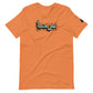 Burnt orange t-shirt featuring central turquoise Arabic calligraphy with black shadow and scattered ink dots, accompanied by a small black logo patch on the sleeve, displayed against a white background.