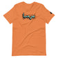 Burnt orange t-shirt featuring central turquoise Arabic calligraphy with black shadow and scattered ink dots, accompanied by a small black logo patch on the sleeve, displayed against a white background.