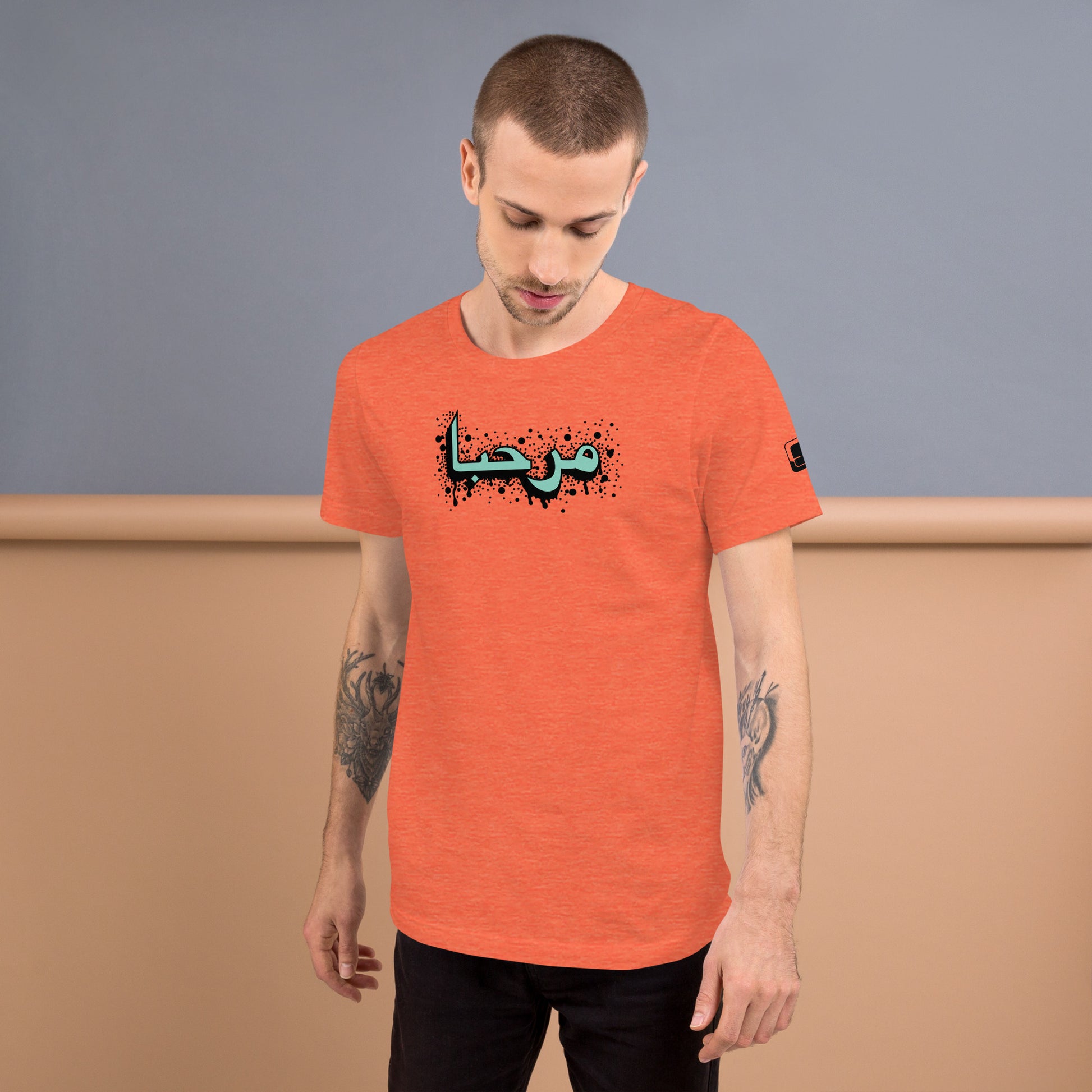 Man with tattoos on his arms looking down, wearing a heather orange t-shirt with a central turquoise Arabic calligraphy design accented with  black splattered ink details, standing against a blue and peach color wall wall. 