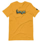 Mustard yellow t-shirt featuring central turquoise Arabic calligraphy with black shadow and scattered ink dots, accompanied by a small black logo patch on the sleeve, displayed against a white background.