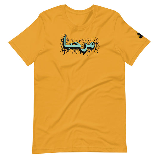 Mustard yellow t-shirt featuring central turquoise Arabic calligraphy with black shadow and scattered ink dots, accompanied by a small black logo patch on the sleeve, displayed against a white background.
