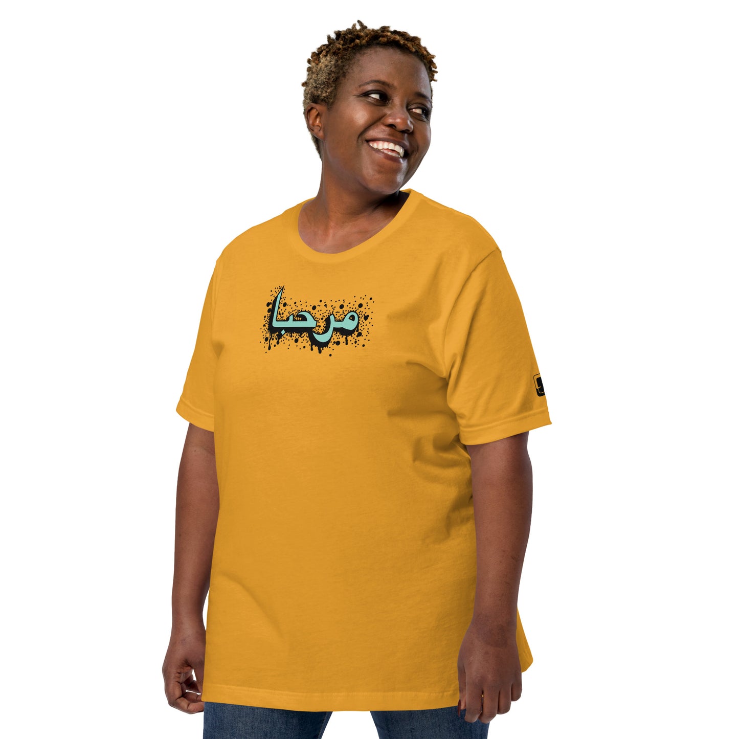 Woman with a joyful smile, sporting a short curly hairstyle, wearing a mustard yellow t-shirt with turquoise Arabic calligraphy and black accents, complete with a small logo patch on the sleeve, set against a white background.