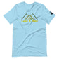 
A light blue t-shirt with a design featuring stylized green mountains above the phrase "Take A Hike" in yellow, bold letters. The mountains are depicted in a simple, modern style with overlapping triangular shapes. The t-shirt has a casual fit with a round neckline and short sleeves, and there's a small black logo patch on the right sleeve. The shirt is laid flat on a white background, emphasizing the clean, bright design and relaxed style of the garment.