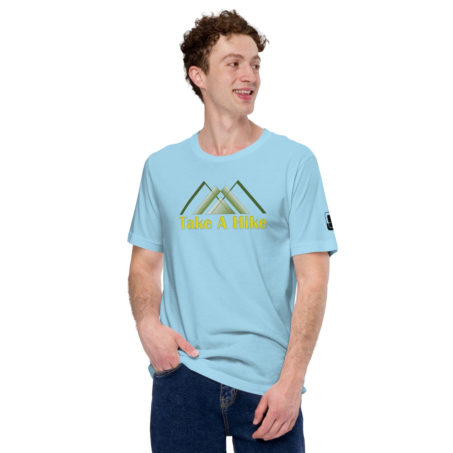 A light-hearted Caucasian man with curly hair is featured wearing a pale blue t-shirt adorned with a green mountain design above the phrase "Take A Hike" in yellow lettering. He is glancing to the side with a smile, his left hand casually in his pocket. The shirt also has a small logo patch on the sleeve, complementing his relaxed style. He stands against a clear, neutral background, enhancing the visibility of the shirt’s design.