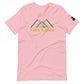A pale pink t-shirt with a design featuring three stylized green mountain peaks above the phrase "Take A Hike" in bold yellow letters. The t-shirt has a crew neckline, short sleeves, and a small black logo patch on the right sleeve. The graphic is centered on the chest, offering a simple yet striking visual theme appropriate for outdoor enthusiasts. The shirt's soft color palette gives it a gentle and appealing look.