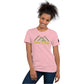 A cheerful African American woman with a beaming smile wears a light pink t-shirt that reads "Take A Hike" in yellow letters, below a stylized green mountain design. She poses casually, leaning slightly to her left, with her left hand resting on her hip. The t-shirt fits comfortably and features a small logo patch on the sleeve. She stands against a plain white background, highlighting her joyful expression and casual style.