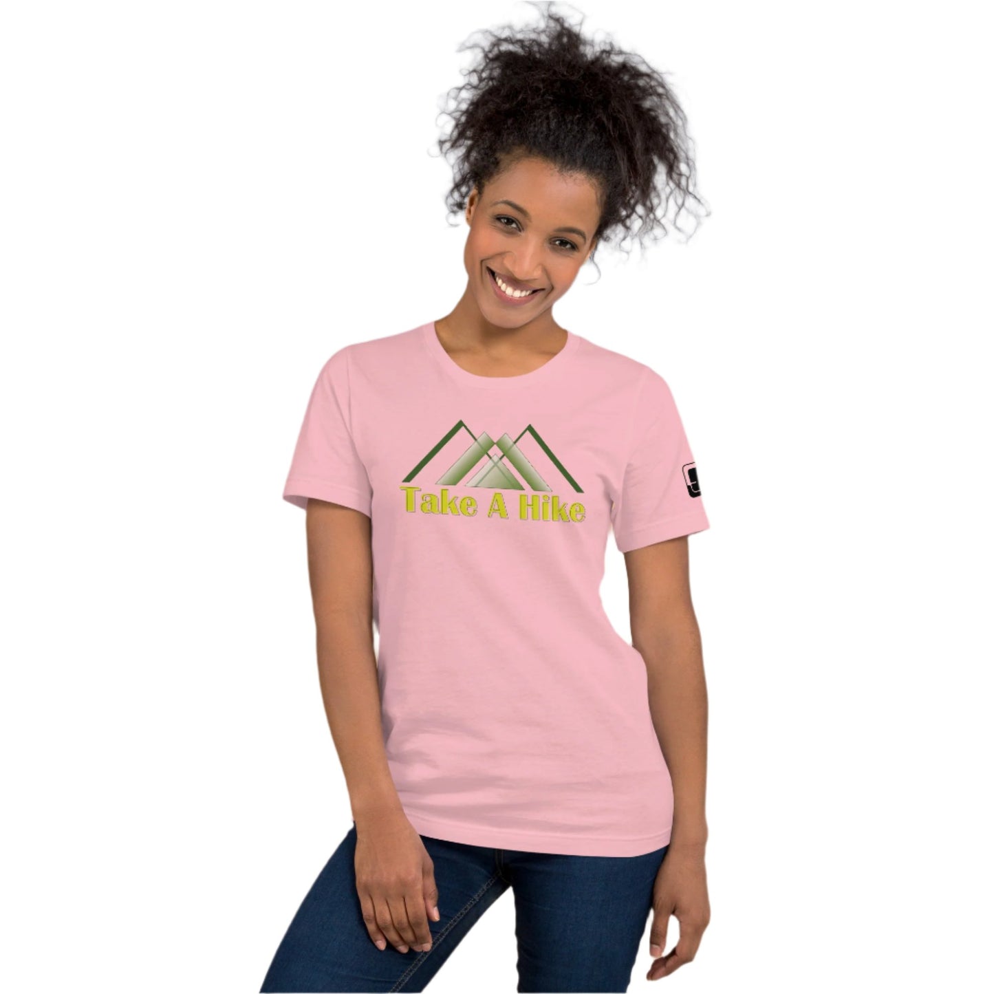 A cheerful African American woman with a beaming smile wears a light pink t-shirt that reads "Take A Hike" in yellow letters, below a stylized green mountain design. She poses casually, leaning slightly to her left, with her left hand resting on her hip. The t-shirt fits comfortably and features a small logo patch on the sleeve. She stands against a plain white background, highlighting her joyful expression and casual style.