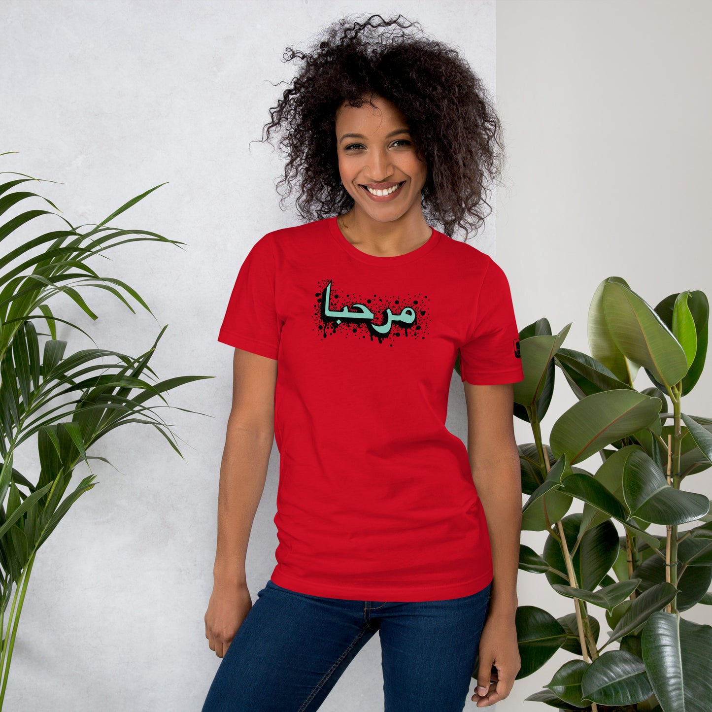Smiling woman with curly hair wearing a red t-shirt featuring a central turquoise Arabic calligraphy design with black accents, a logo patch on the sleeve, styled with blue jeans, standing in an indoor setting with green plants in the background.
