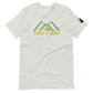 A light grey t-shirt featuring a stylized design of green mountains above the phrase "Take A Hike" in bold, yellow text. The graphic includes three overlapping mountain peaks, creating a clean, geometric look. The t-shirt has a crew neck and short sleeves with a small black logo patch on the right sleeve. The design is showcased against a white background, emphasizing the shirt's outdoor-themed graphic.