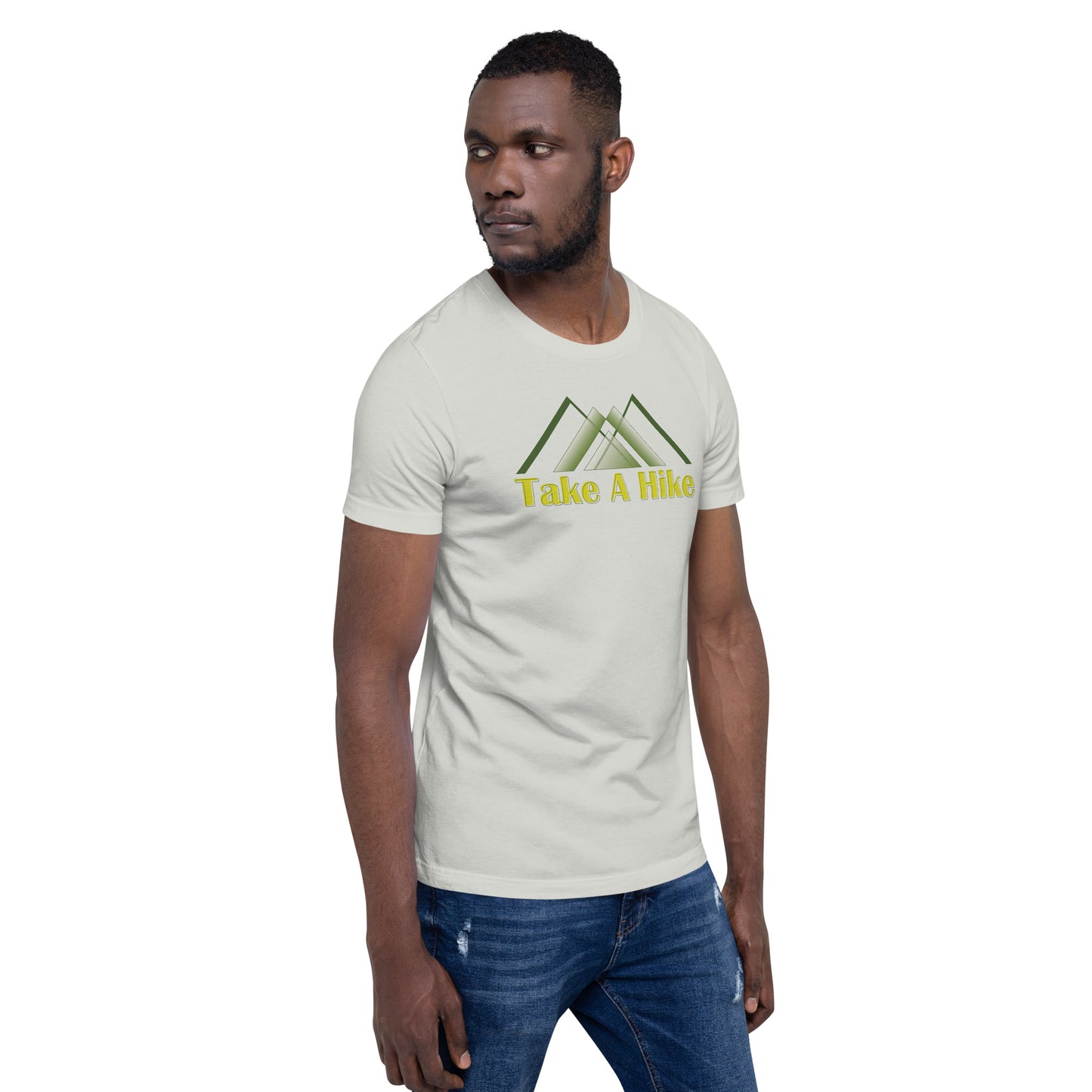 A somber African American man with a focused expression wears a light beige t-shirt featuring a stylized green mountain design above the phrase "Take A Hike" in yellow text. He is gazing off to the side with a serious look. The fitted t-shirt complements his lean physique, and he pairs it with dark blue jeans. The backdrop is neutral, emphasizing his overall attire.