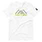 A plain white t-shirt with a design featuring stylized green mountains above the phrase "Take A Hike" in yellow, bold letters. The mountains are depicted in a simple, modern style with overlapping triangular shapes. The t-shirt has a casual fit with a round neckline and short sleeves, and there's a small black logo patch on the right sleeve. The shirt is laid flat on a white background, highlighting the vibrant design and clean lines of the garment.