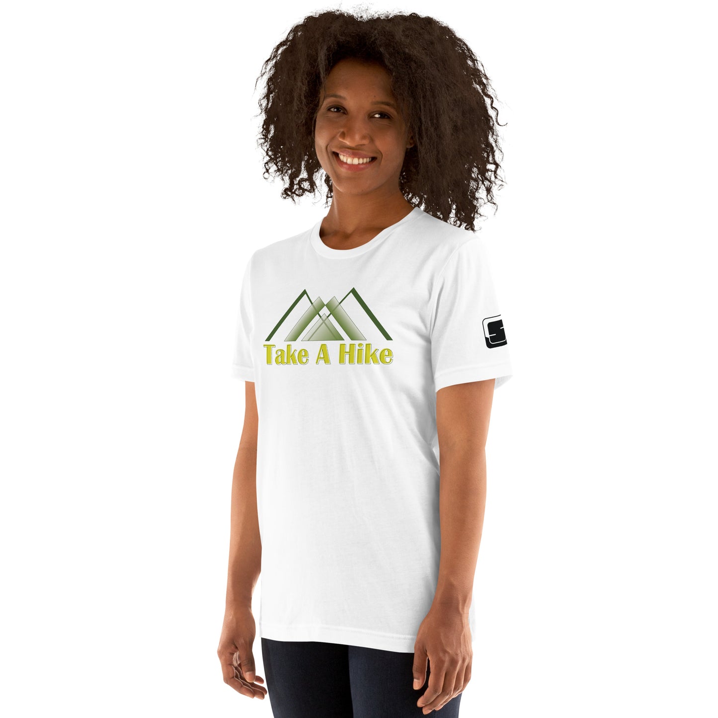 A cheerful African American woman with voluminous curly hair wears a white t-shirt featuring stylized green mountains and yellow text that states “Take A Hike”. She stands confidently, smiling at the camera, her hands slightly behind her hips. The t-shirt's design is clearly visible on her chest, and she has a relaxed posture in front of a white background. A small logo patch is visible on the sleeve of the t-shirt, adding a detail to the simple outdoor-themed attire.